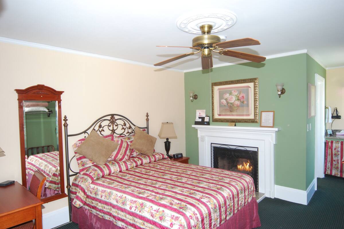 Um quarto com cama, ventilador de teto, lareira, espelho e mesa de cabeceira.