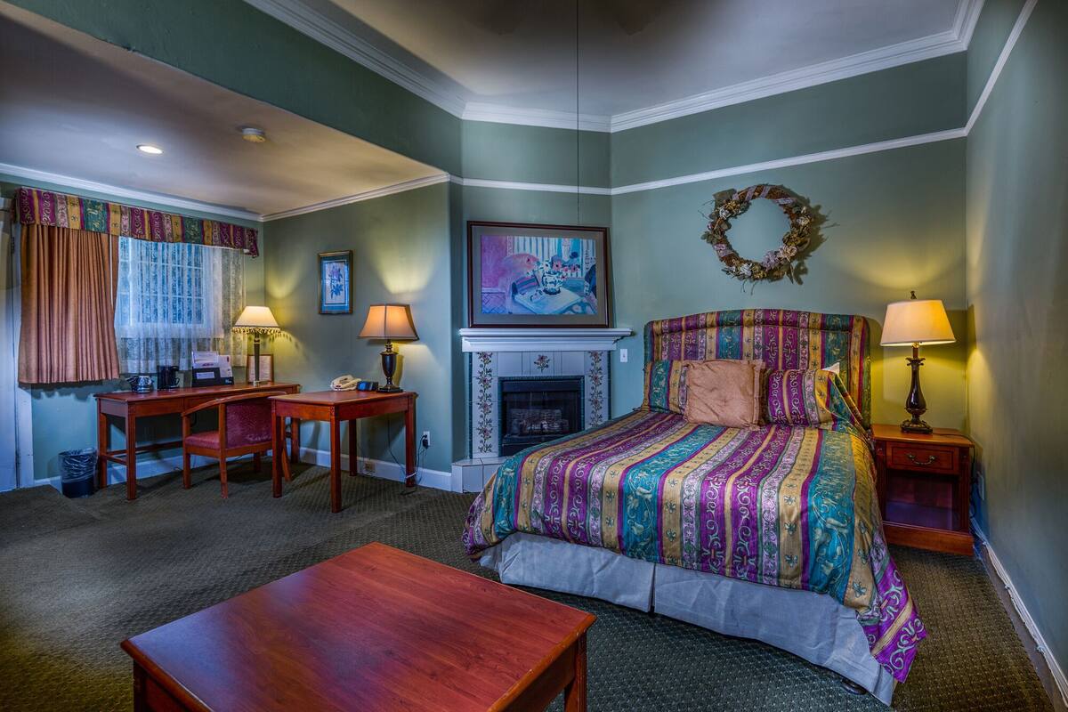Quarto de hotel com uma cama, duas mesas de cabeceira, uma escrivaninha com cadeira, lareira e janela com cortinas.