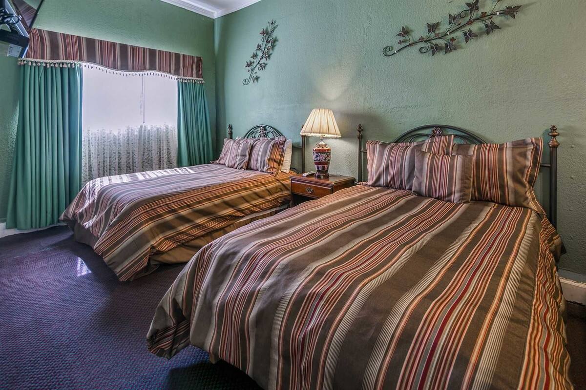 Quarto com duas camas, janela com cortinas, luminária e detalhes decorativos na parede.