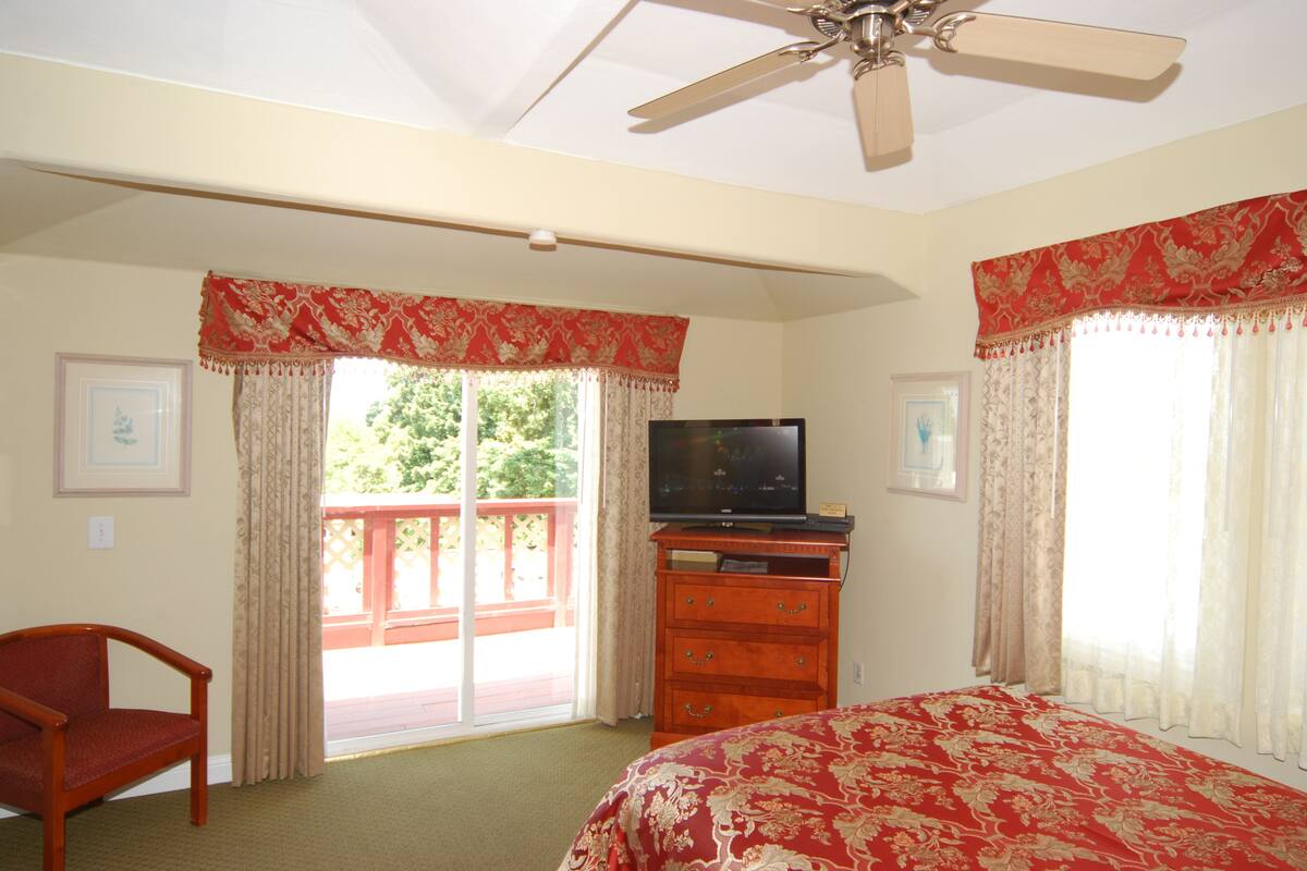Quarto com cama, televisão sobre uma cômoda de madeira, ventilador de teto e varanda com grades vermelhas.
