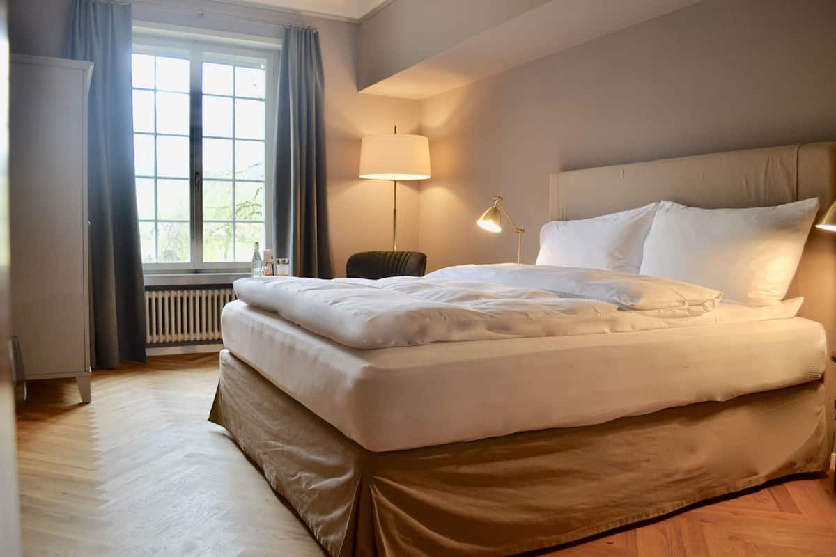 Una cama bien hecha con ropa blanca, una lámpara de noche y vistas a una ventana con cortinas.
