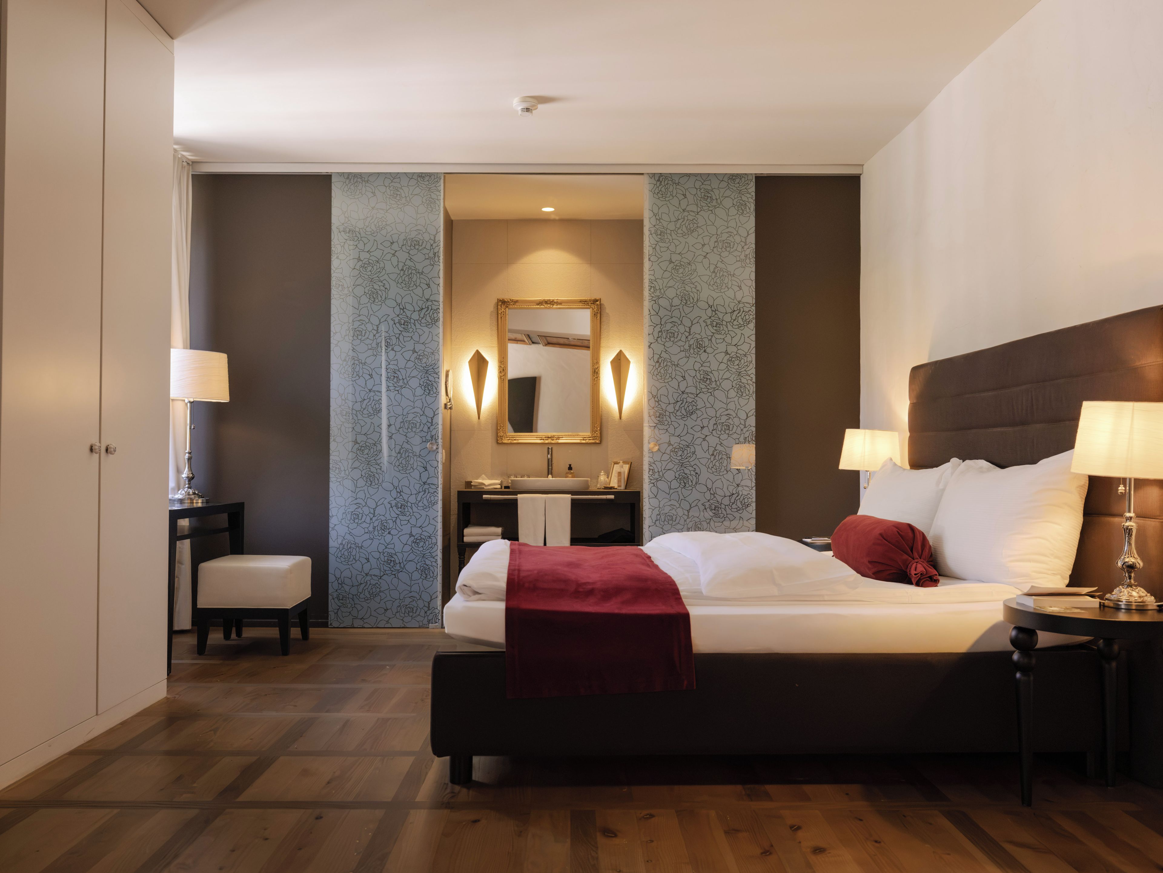A modern hotel room with a large bed, bedside tables with lamps, a vanity with a mirror, and a wooden floor.