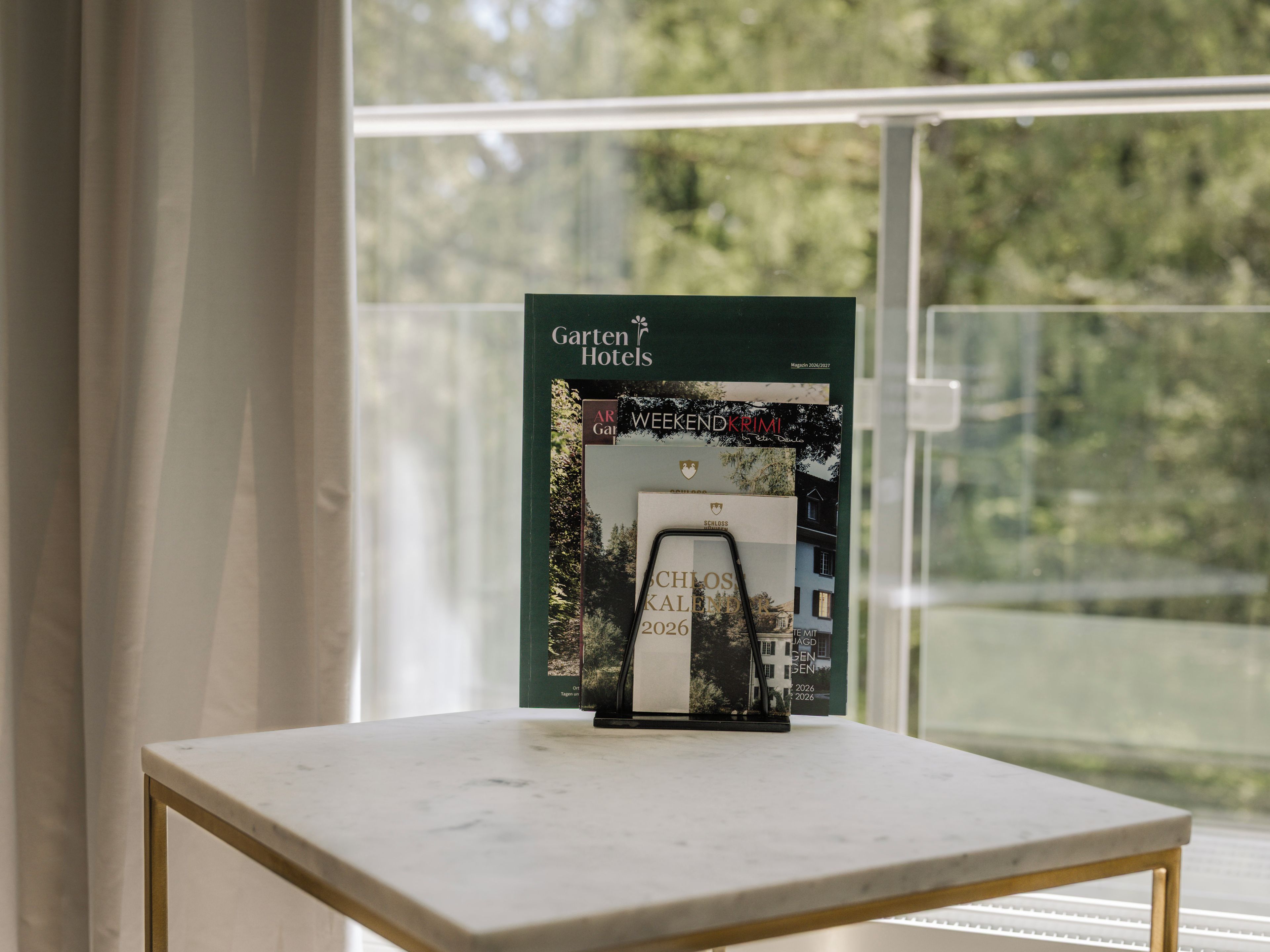 Un folleto con el título 'Garten Hotels' sobre una mesa cerca de una ventana con vistas a árboles.