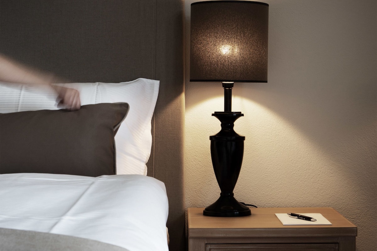 Una cama bien hecha con ropa de cama blanca y marrón, una mesita de noche con una lámpara y un cajón.