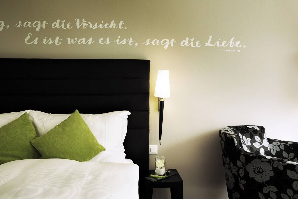 Una cama bien hecha con almohadas verdes, una mesita de noche con lámpara y un sillón floral.