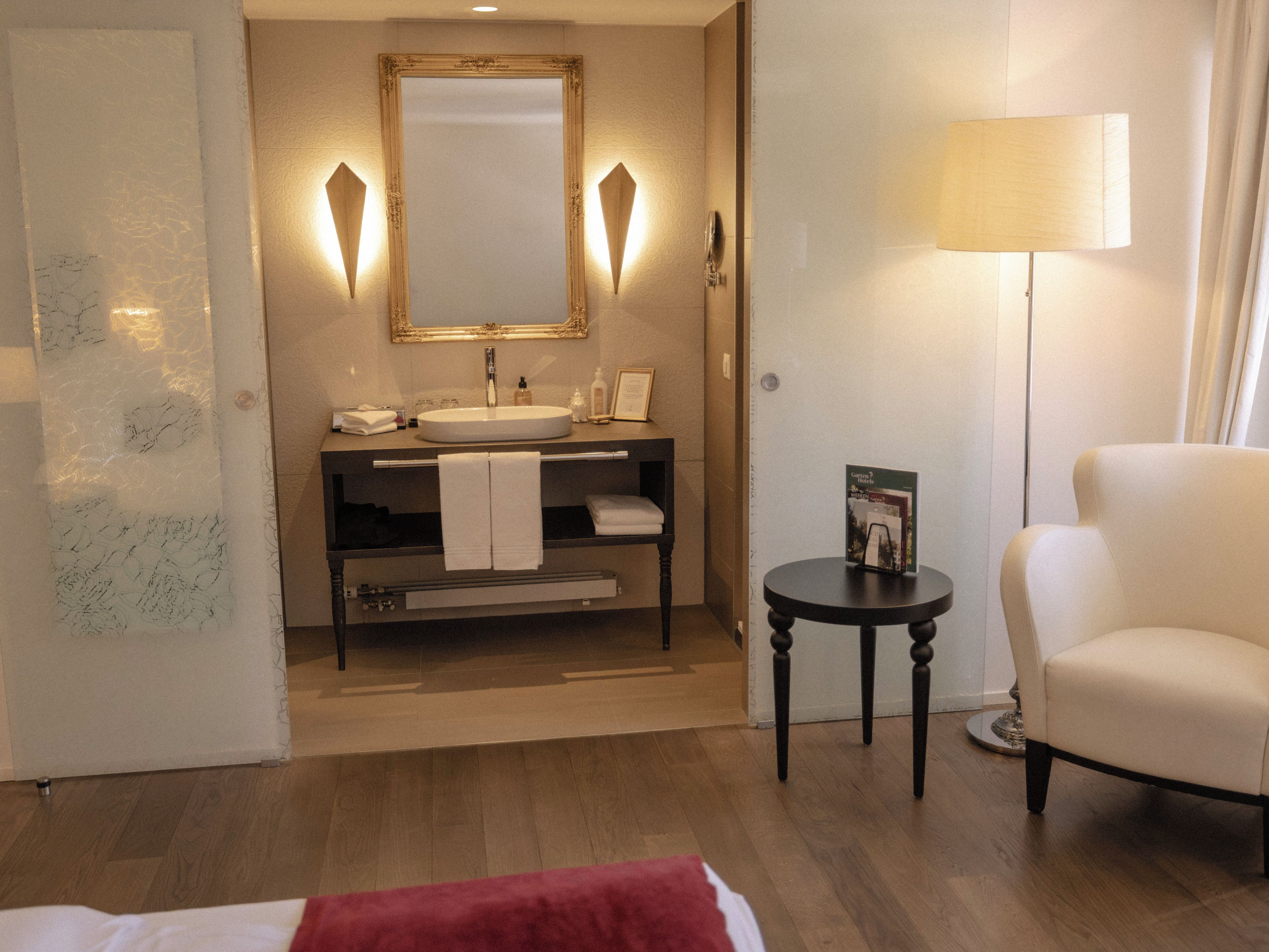 Un baño de hotel con un espejo grande, un lavamanos, un perchero para toallas, una mesita con una lámpara y una silla.