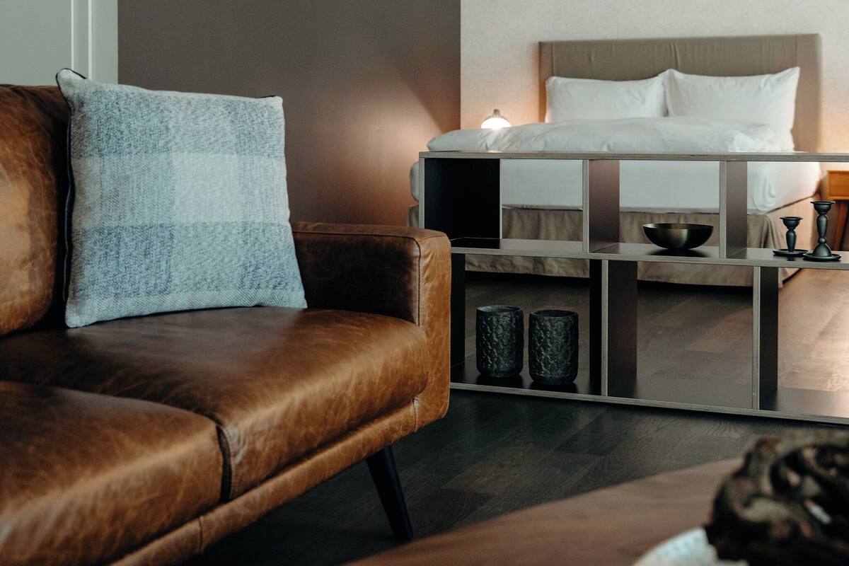 Una habitación de hotel moderna con sofá de cuero, una mesita de madera, cama con ropa blanca y un diseño minimalista.