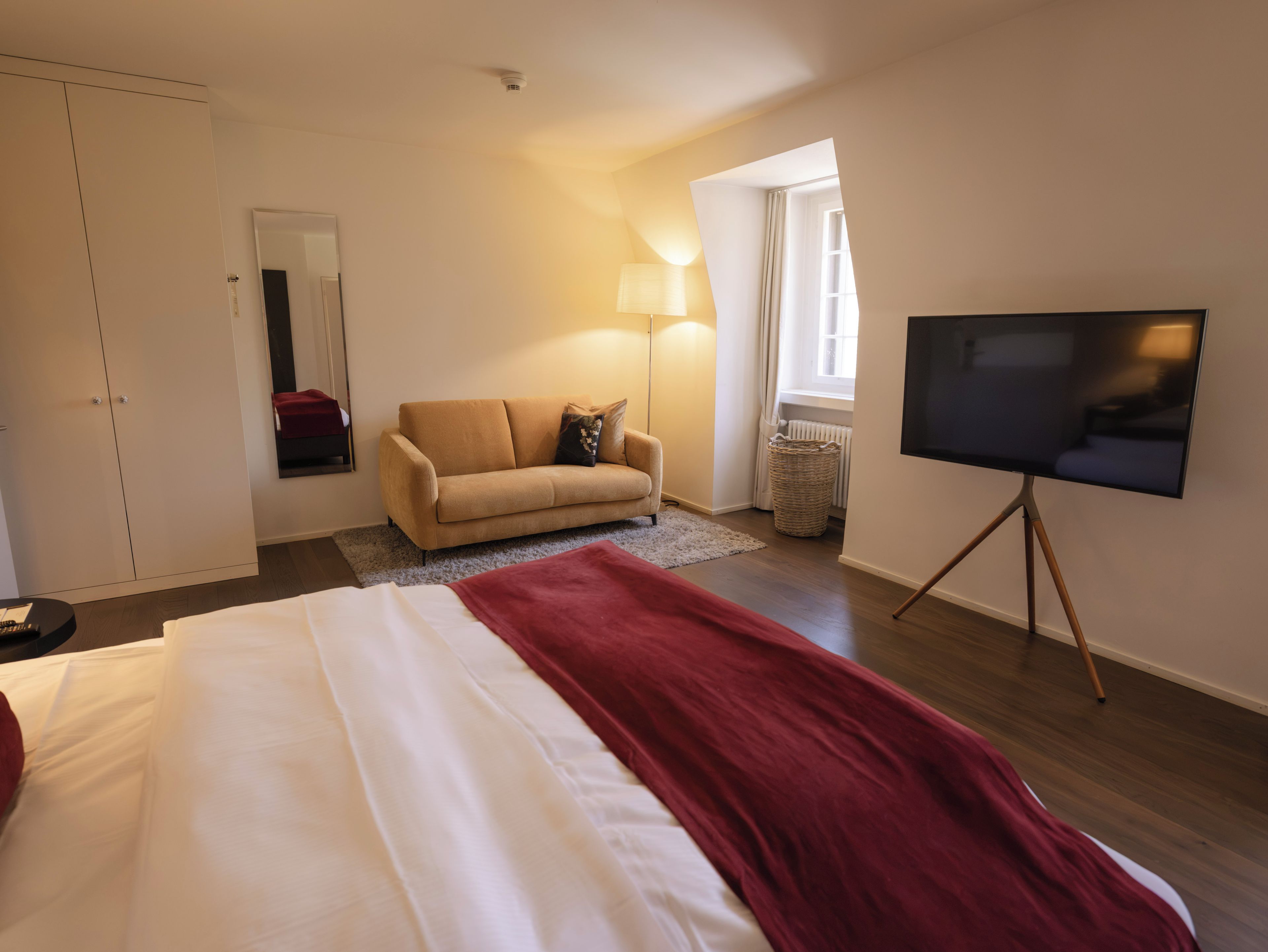 Ein Hotelzimmer mit Bett, einer Couch, einem Fernseher auf einem Schrank, einer Lampe und einem Fenster mit Vorhängen.