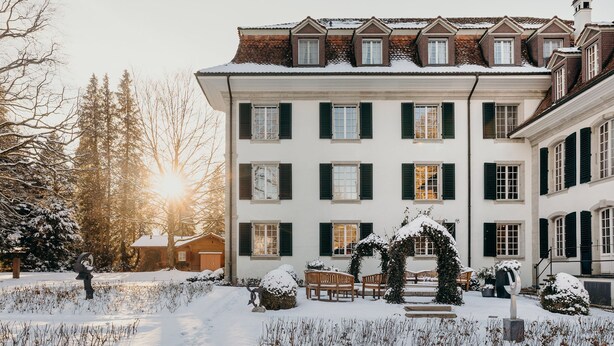 A large, multi-story building with a symmetrical facade, multiple windows with shutters, and a snow-covered garden with benches. - Schloss Huenigen (Konolfingen)