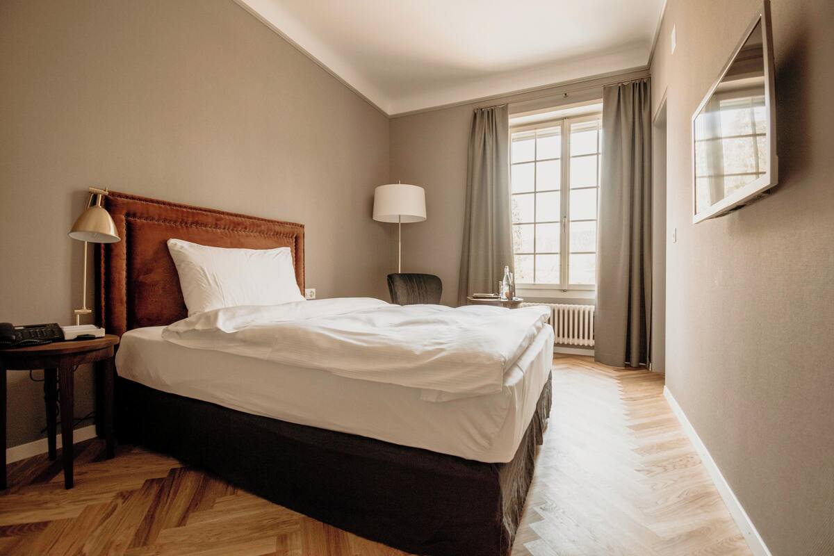 Una habitación de hotel con una cama grande, una mesita de noche con lámpara, una silla, una ventana con cortinas y un radiador.