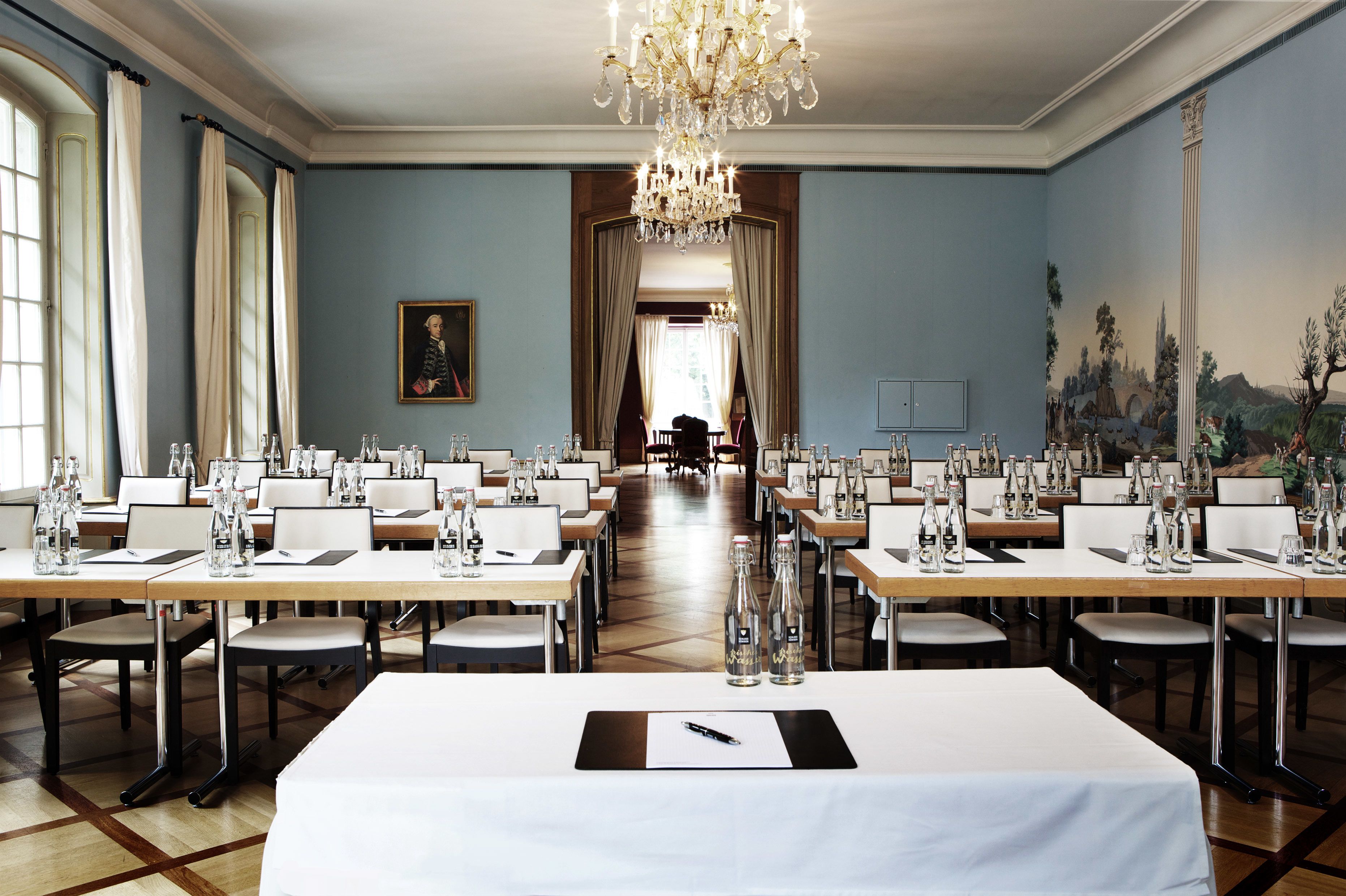Uma sala de jantar formal com longas mesas preparadas para refeição, um candelabro e um quadro grande na parede.
