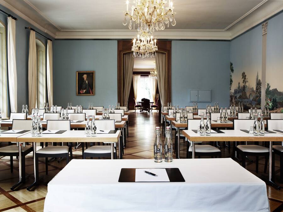 Uma sala de jantar formal com longas mesas preparadas para refeição, um candelabro e um quadro grande na parede.