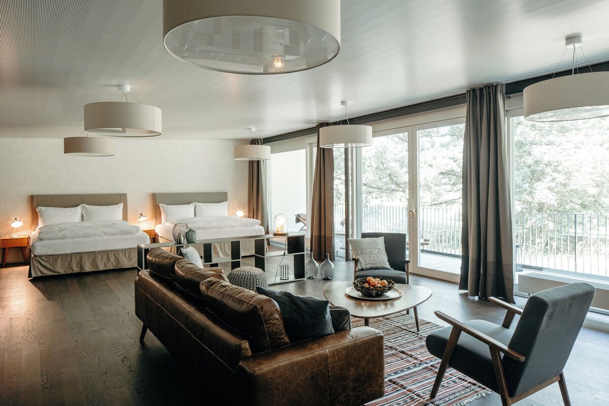 A modern hotel room with a large bed, a sofa, a coffee table, and a balcony with curtains.