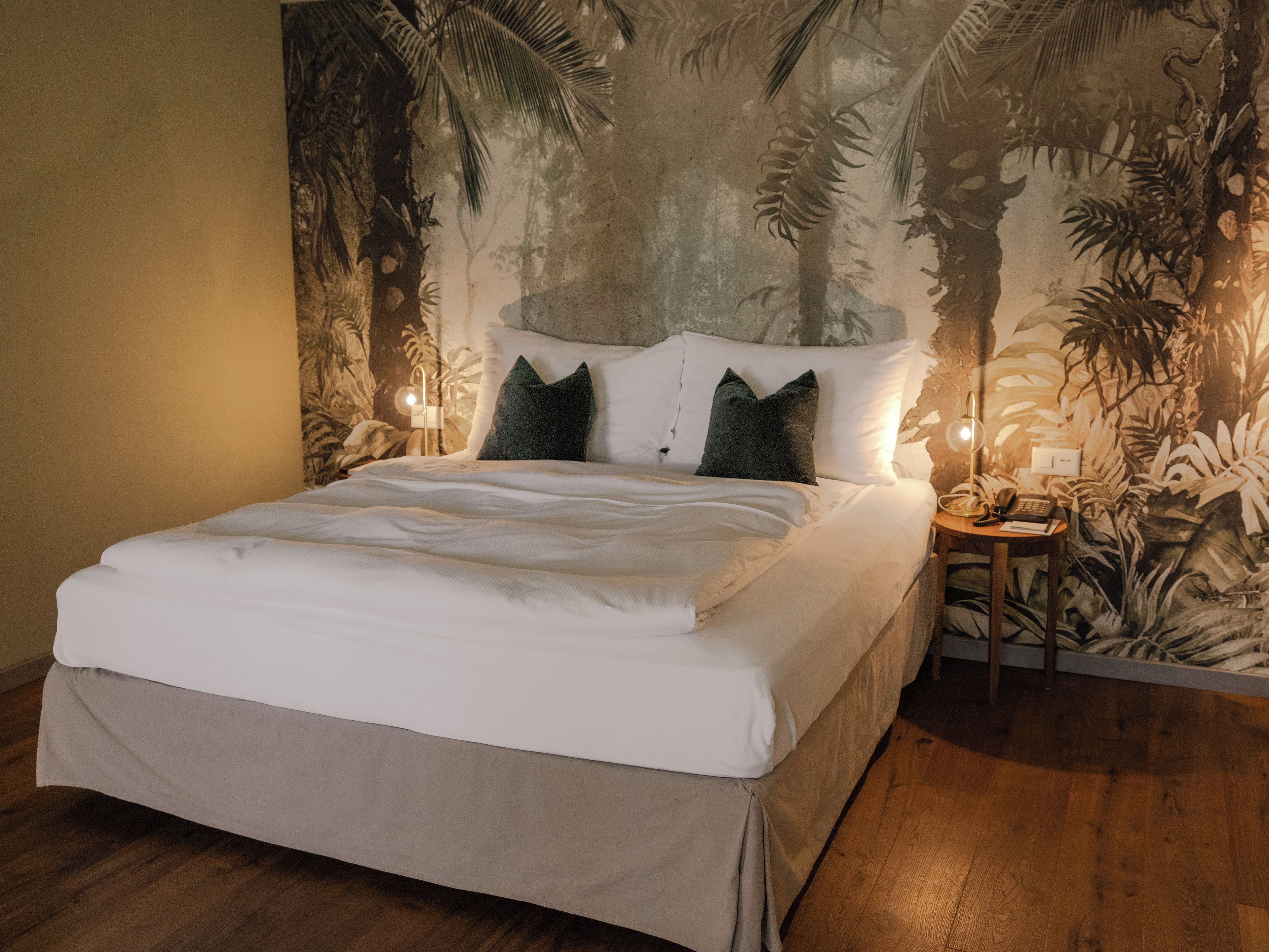 A neatly made bed with white linens and two dark pillows, a nightstand with a lamp, and a wall mural of a forest scene.