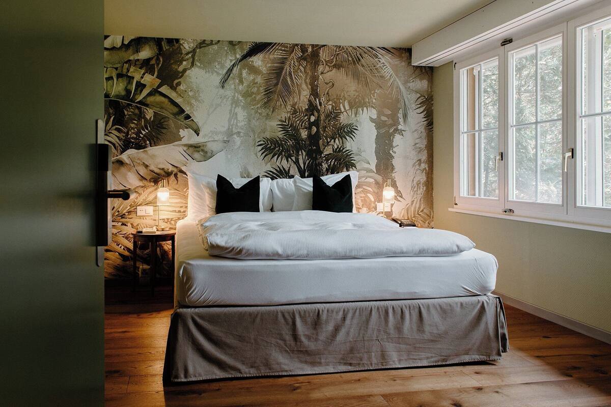 Un dormitorio con cama, mesita de noche, lámpara y ventana con vistas a árboles.
