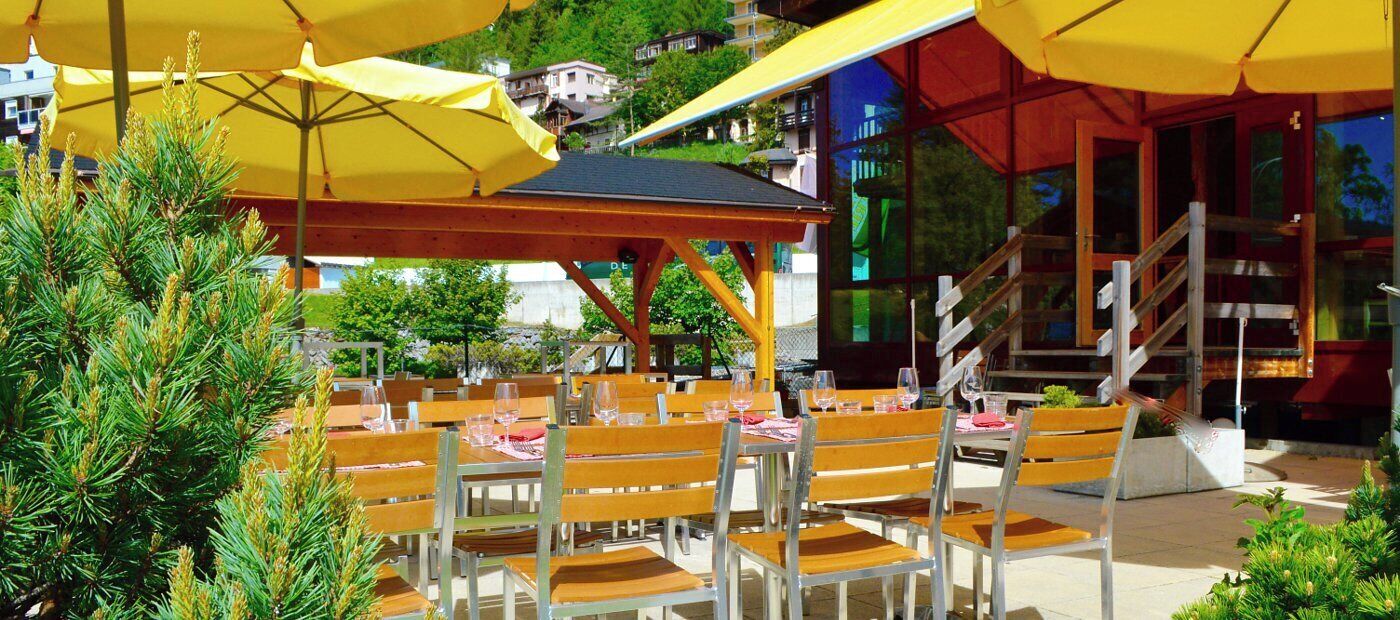 Un área de comedor al aire libre con mesas y sillas de madera, sombrillas amarillas y una escalera que lleva a un edificio con ventanas de vidrio.