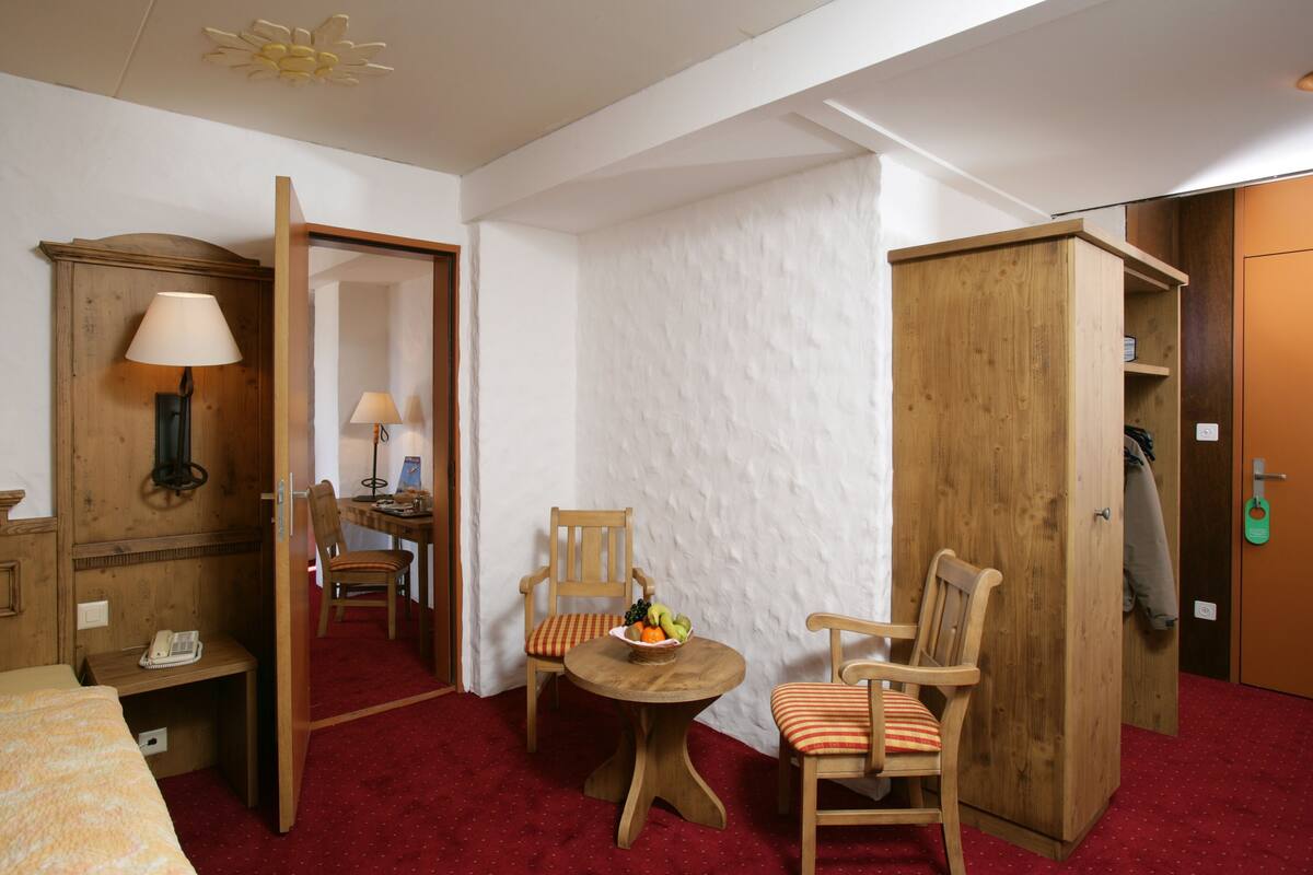 Une chambre d’hôtel avec une armoire en bois, un bureau avec une lampe, une chaise, une petite table ronde avec un panier de fruits et un lit recouvert d’une couverture à motifs.