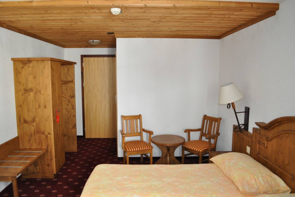 Une chambre d’hôtel avec une armoire en bois, un lit recouvert d’une couvre-lit à motifs, deux chaises en bois disposées autour d’une petite table et une lampe sur la table de chevet.