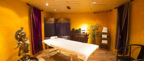 A room with a wooden floor, a massage table, a statue, a shelf with towels, and a bench.