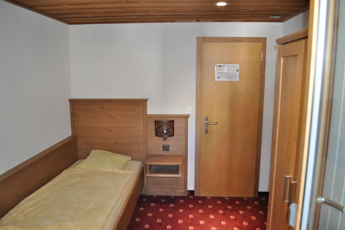 Une petite pièce avec un lit en bois, une porte sur laquelle est affichée une note et un sol à motifs rouges.