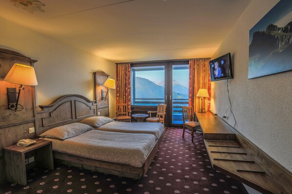 A hotel room with two beds, a TV, a desk, a chair, a lamp, and a view of mountains.
