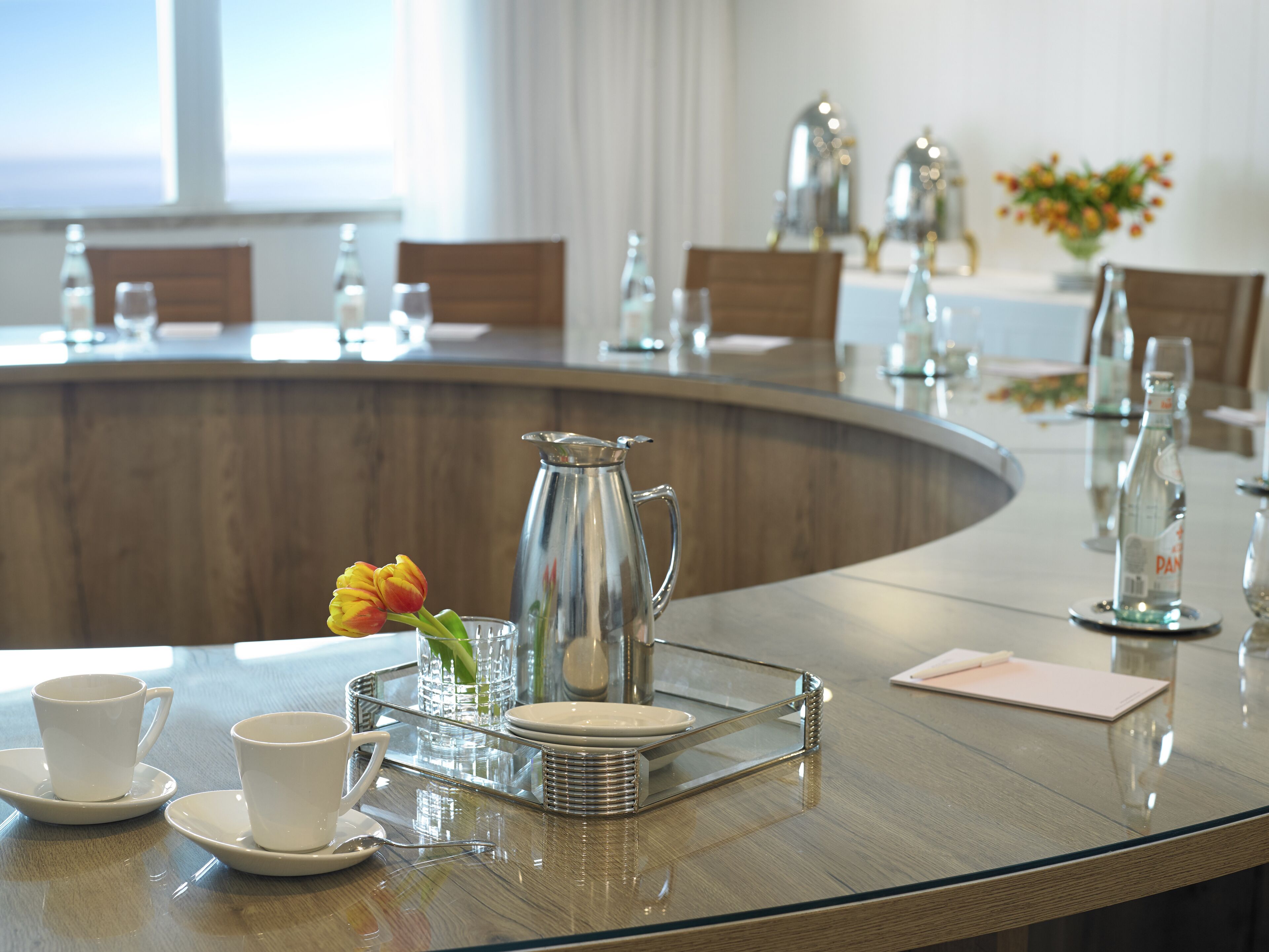 A modern dining area with a curved glass table, white plates, cups, and a pitcher.