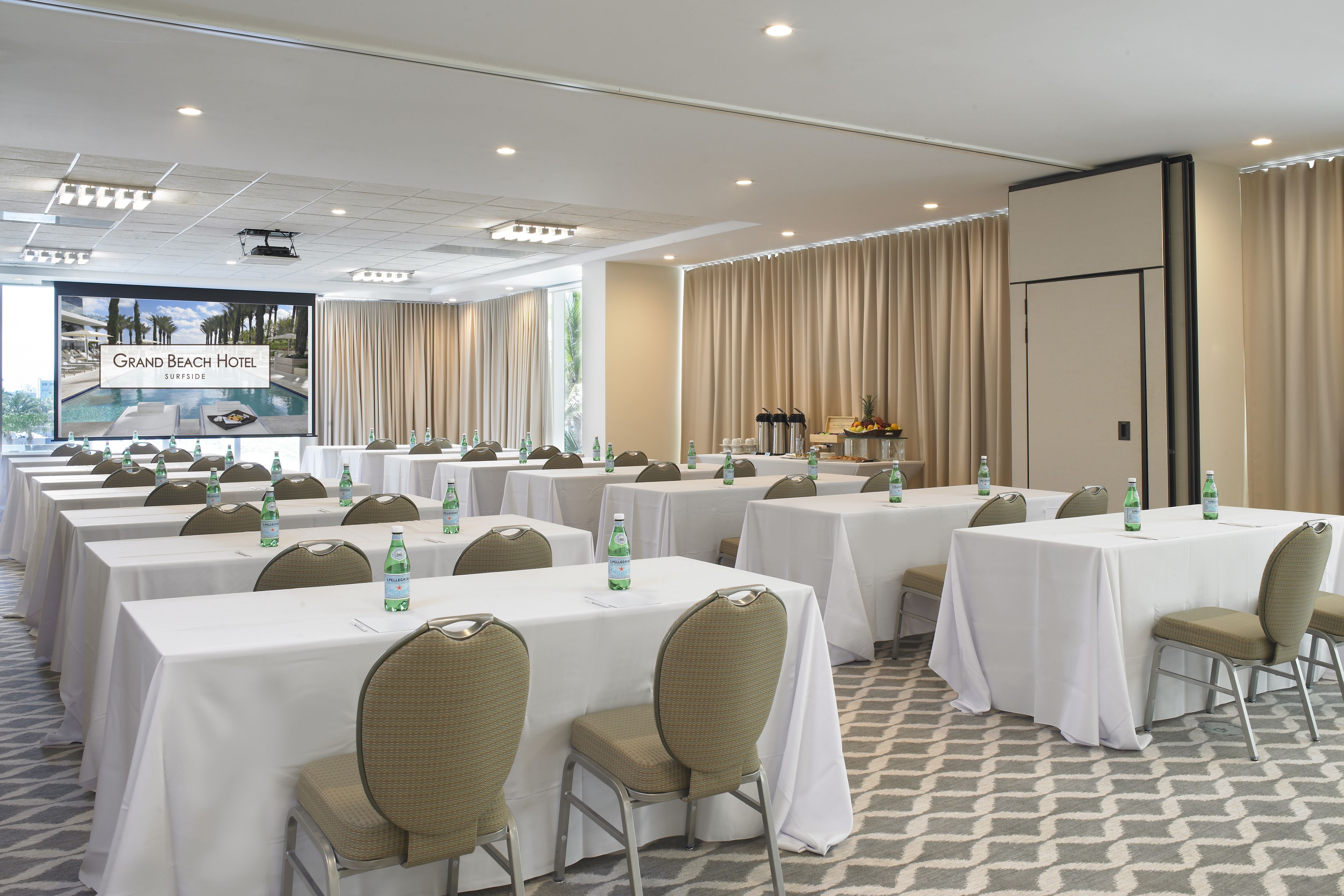A conference room with multiple tables, chairs, and a large screen displaying the Grand Beach Hotel.