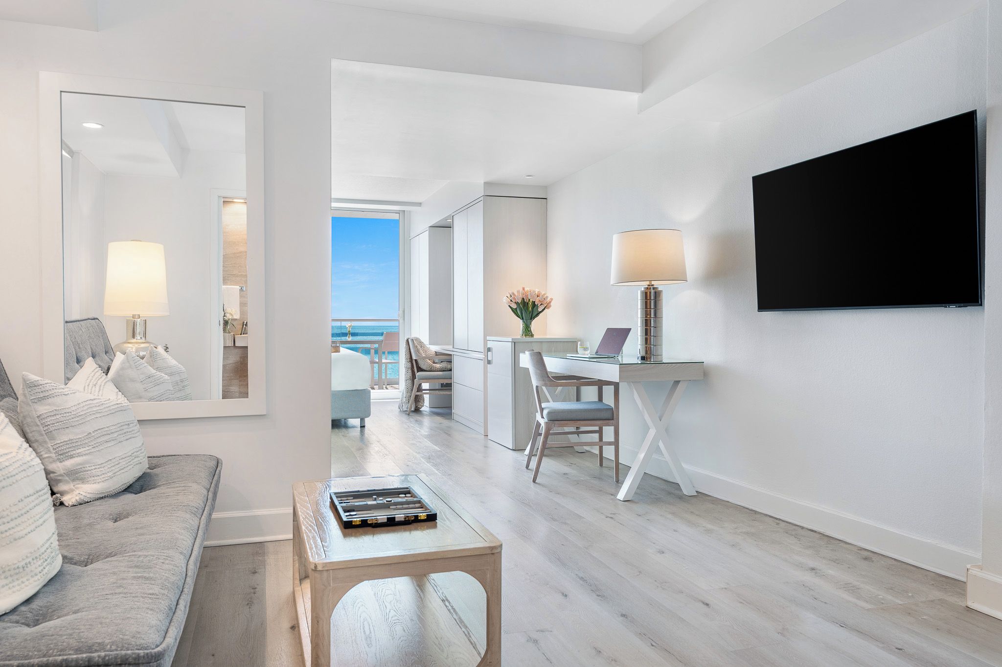 A modern living room with a sofa, a coffee table, a TV mounted on the wall, and a view of the ocean through a glass door.