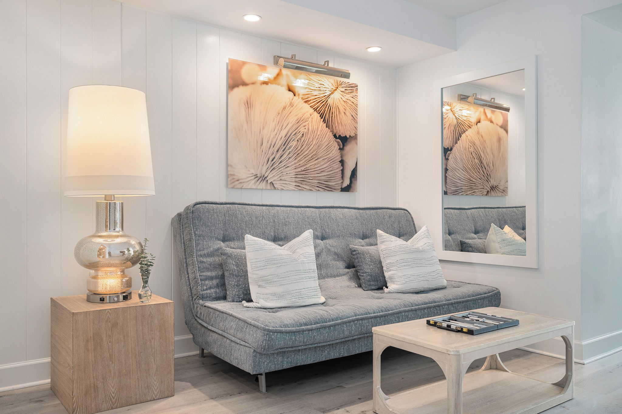 A modern living room with a grey sofa, white cushions, a side table with a lamp, and a coffee table.