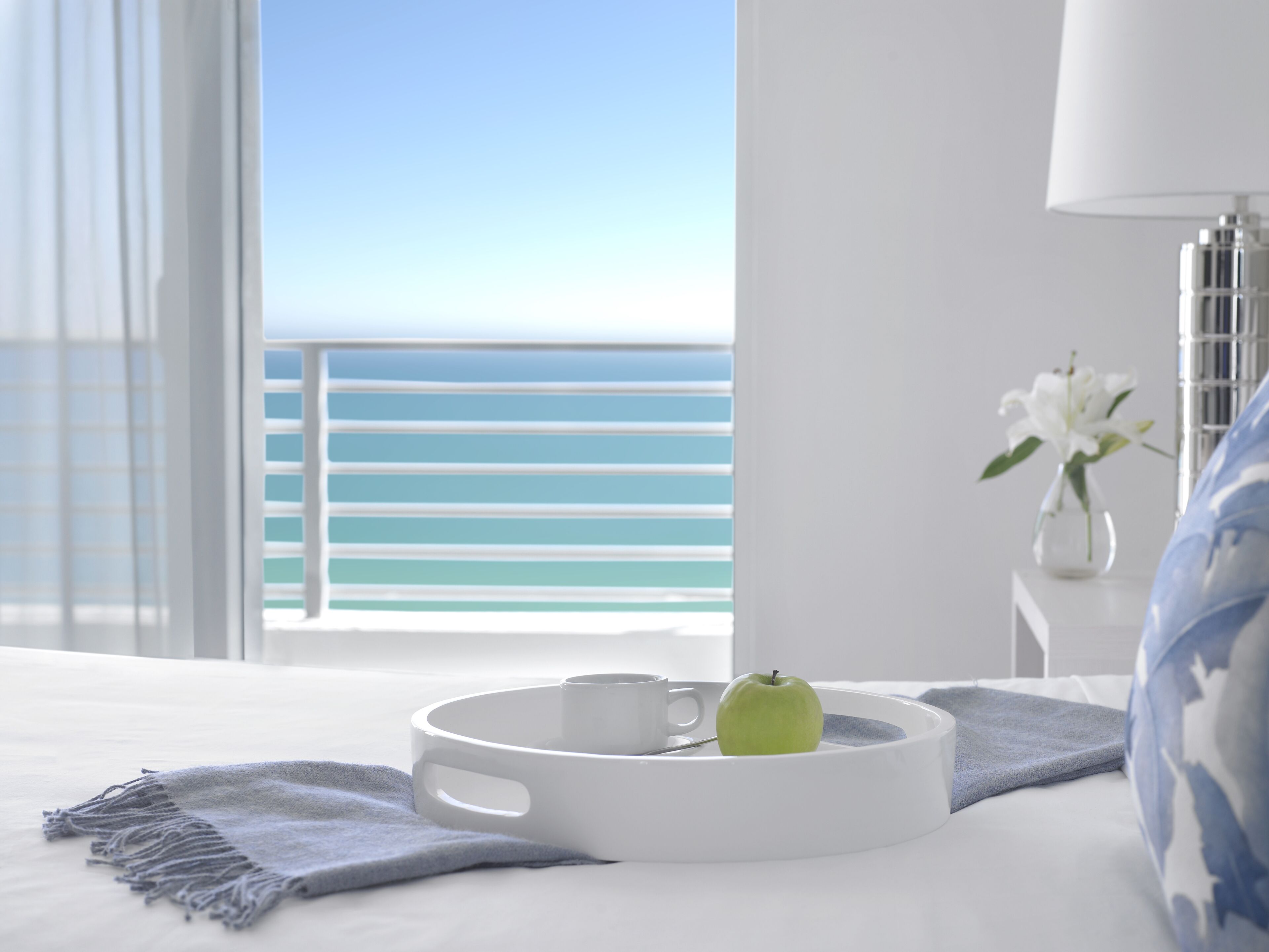 Habitación de hotel con una cama, una mesita de noche con una lámpara, un florero con flores y una bandeja con una manzana verde y una taza blanca.