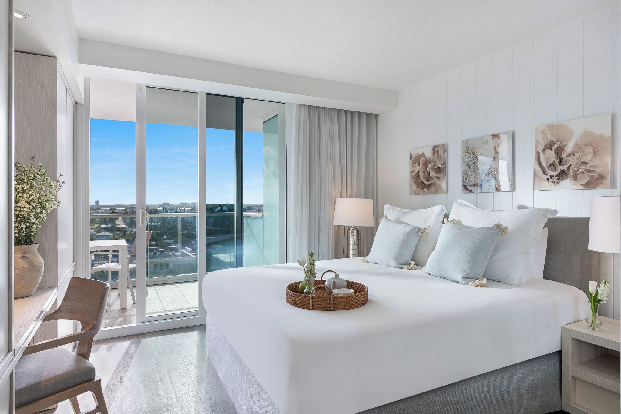 A hotel room with a large bed, a chair, a table, and a view of the city through large windows.
