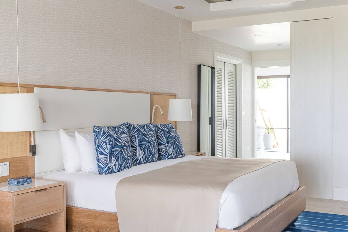 A modern hotel room with a large bed, a wooden headboard, a nightstand, a bedside table, and a window with a view of greenery outside.