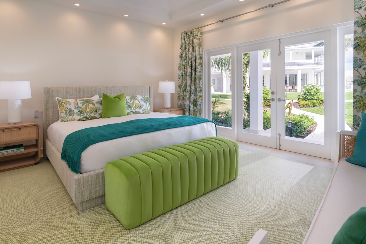 A hotel room with a bed, a bench, a nightstand, a lamp, and a view of the outside.