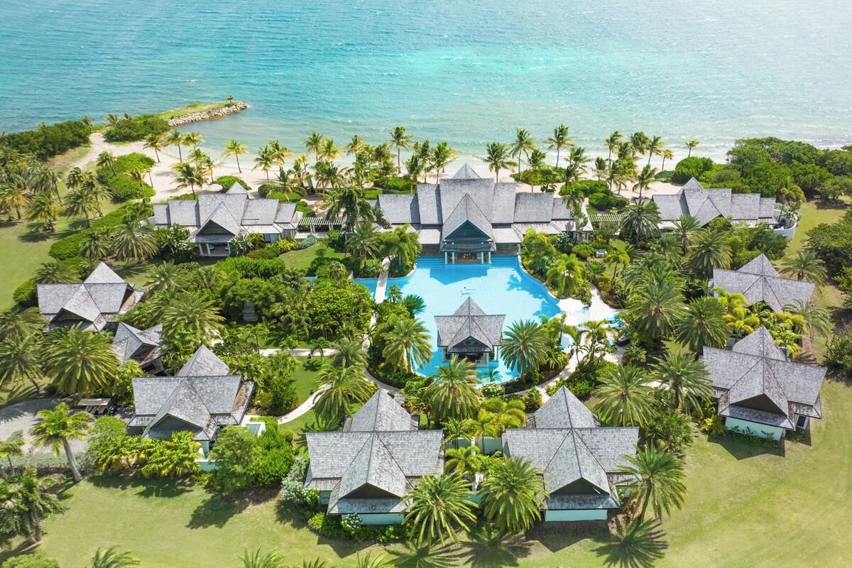 Vista aérea de un resort con piscina central, rodeado de varias cabañas, palmeras y una playa.