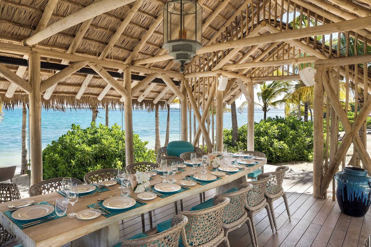 An outdoor dining area with a wooden table set for a meal, surrounded by wicker chairs, and overlooking a body of water.
