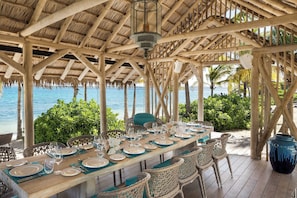 Un espace de restauration en plein air avec une table en bois dressée pour un repas, entourée de chaises en osier, et donnant sur une étendue d’eau.