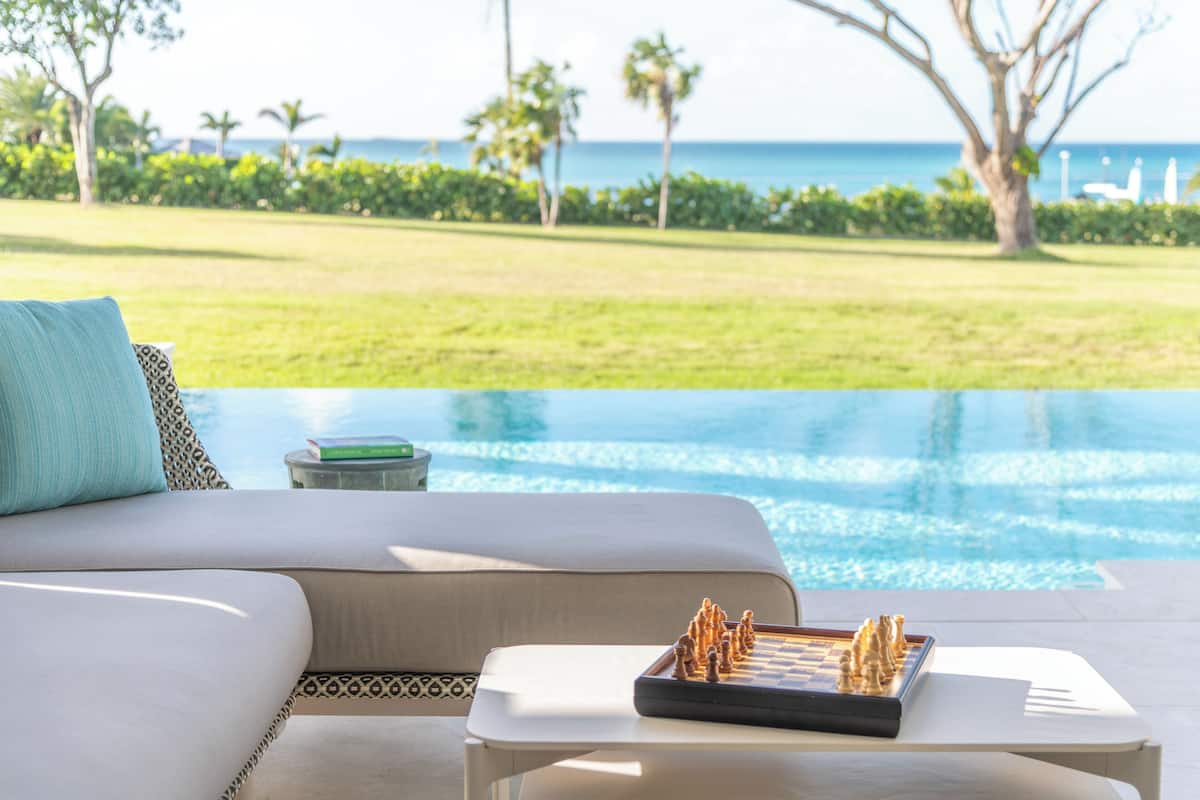 A poolside area with a chessboard on a table, a sofa with a patterned pillow, and a view of the ocean and greenery.