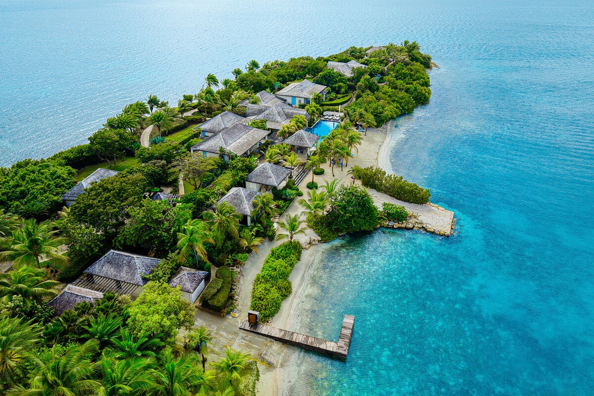 Vista aérea de un resort tropical con varias cabañas, un mar azul cristalino y una playa de arena.
