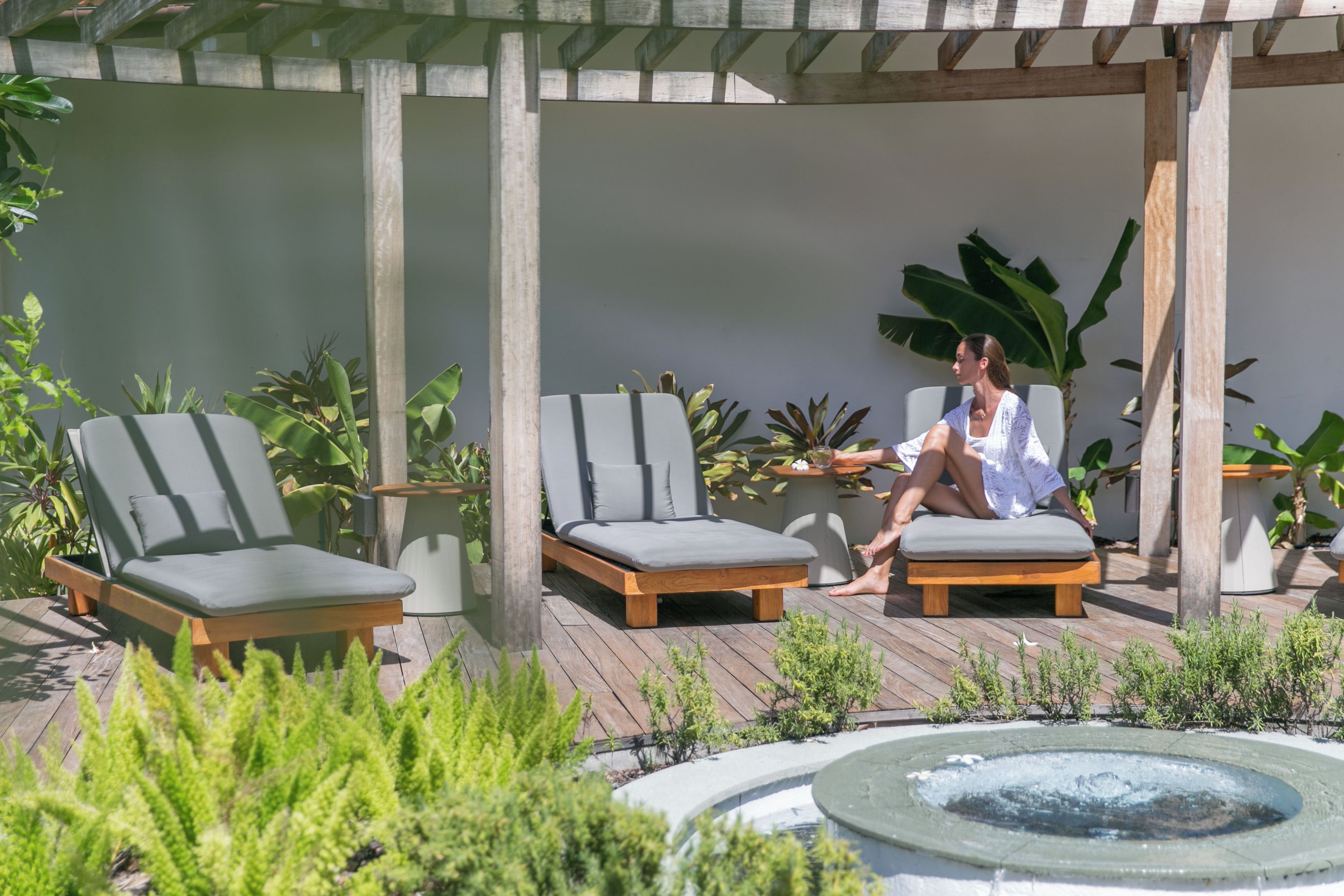 Une personne est assise sur un fauteuil de jardin dans un espace extérieur abrité, doté d’une terrasse en bois, et entourée de plantes en pot et d’une petite fontaine.