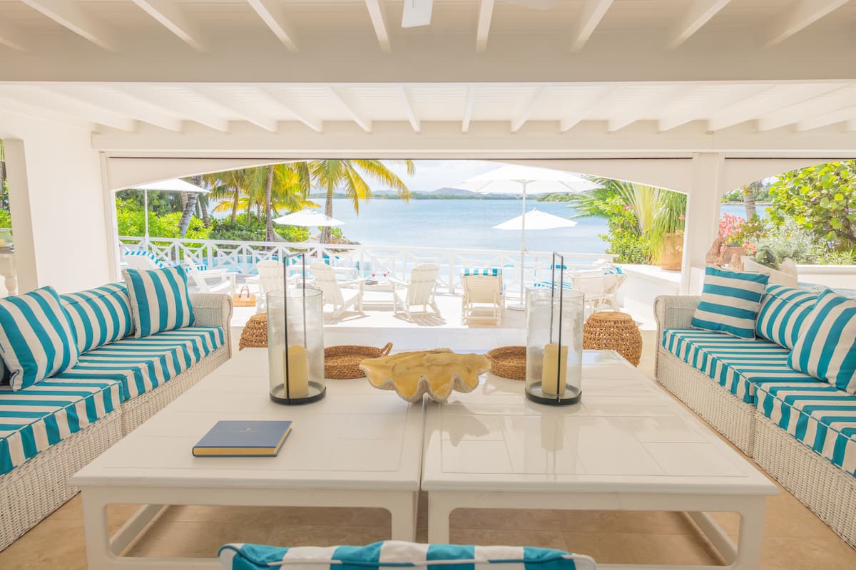 A covered outdoor seating area with white furniture and striped cushions, overlooking a pool and tropical scenery.