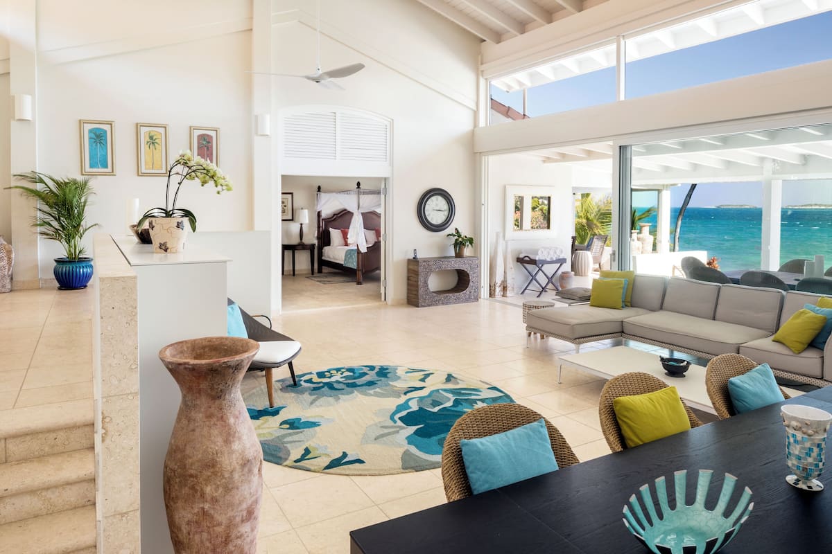 A spacious living area with a large window overlooking the sea, a sofa set, a dining table, and a staircase leading to an upper level.