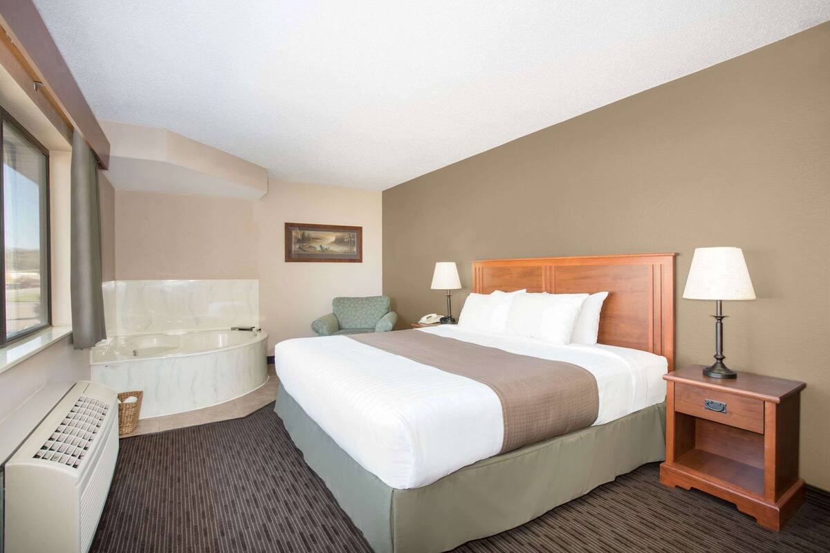 A hotel room with a bed, a desk, a chair, a lamp, a phone, a painting, and a window.