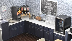 Cocina con armarios azul oscuro, una estación de café, una pizarra con dibujos relacionados con el desayuno y un letrero que indica los horarios de desayuno gratuito.