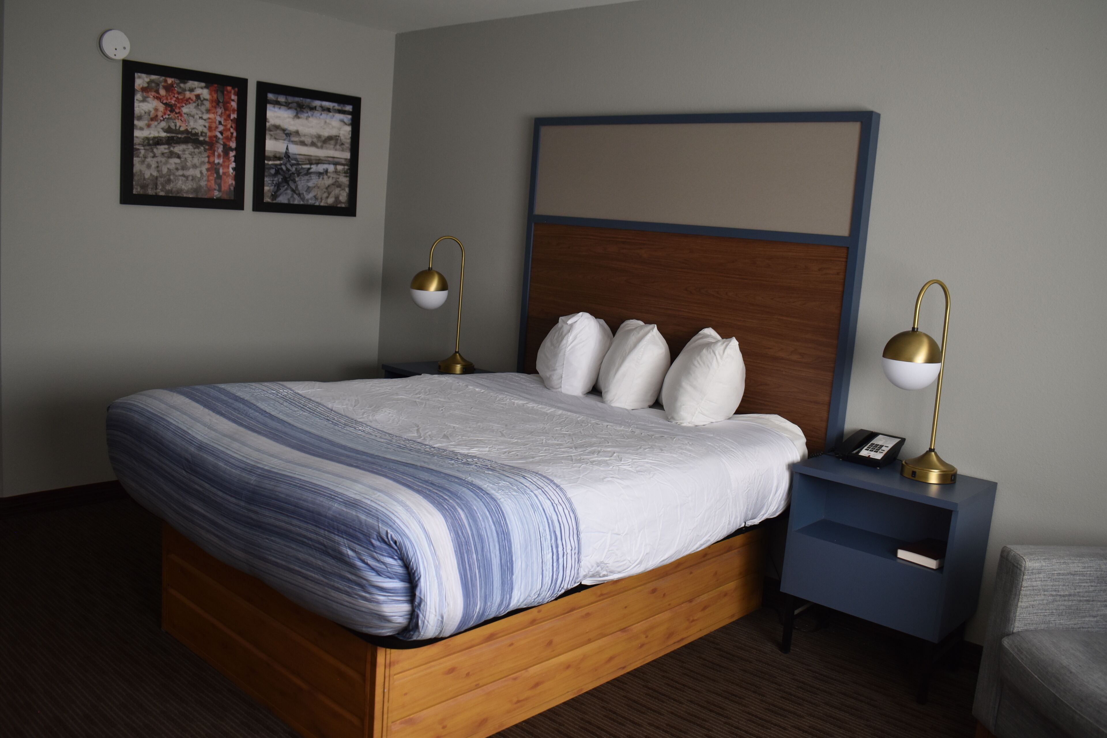 A neatly made bed with a striped blanket, a wooden headboard, and two bedside lamps. There are two framed pictures on the wall and a small bedside table with a phone.