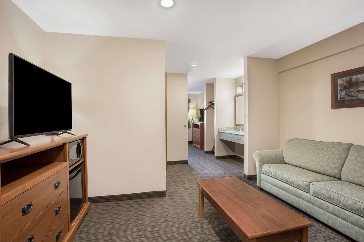 A hotel room with a television on a wooden stand, a sofa, a wooden coffee table, and a kitchen area in the background.
