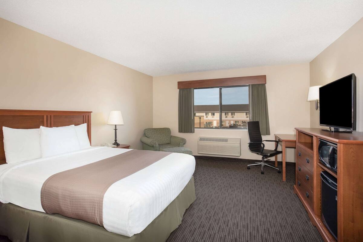 A hotel room with a large bed, a desk with a chair, a television, and a view of buildings through the window.