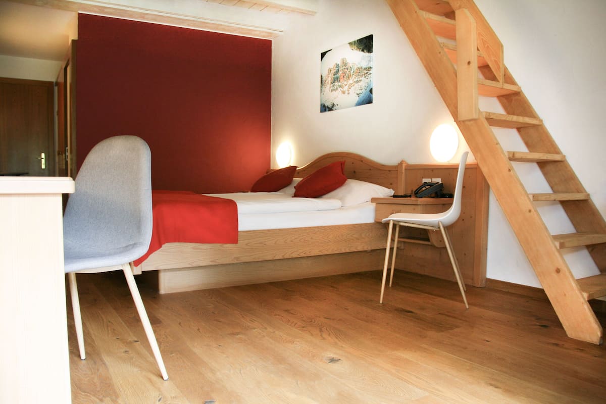 A single bed with a wooden headboard, a desk with a chair, a red accent wall, a wooden ladder, and a painting on the wall.