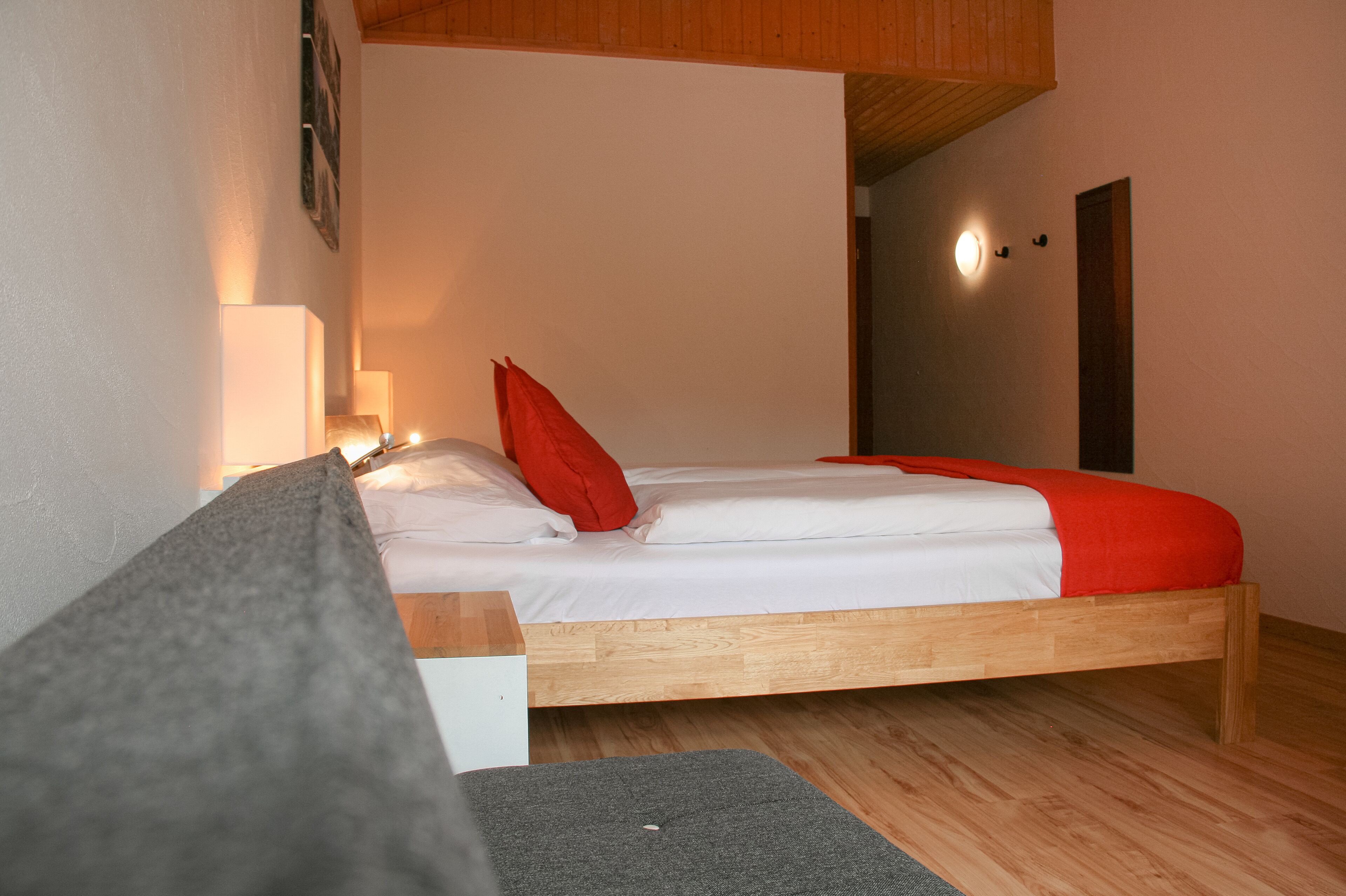 A single bed with a wooden frame, white bedding, and a red pillow. There is a bedside lamp and a small table with a grey cushion. The room has wooden flooring and a sloped ceiling.