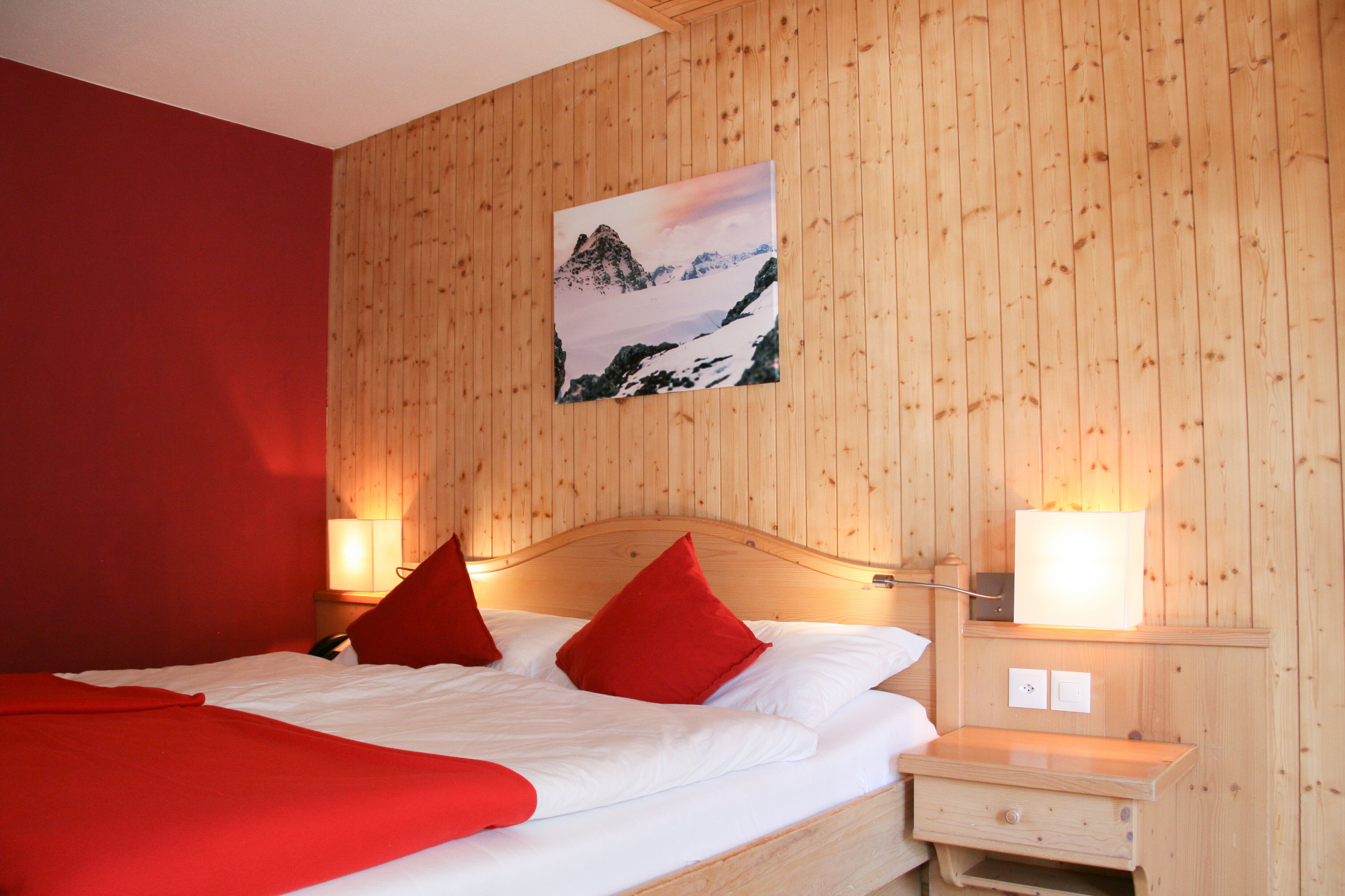 A room with a wooden paneled wall, a bed with red pillows, and a nightstand with a lamp.