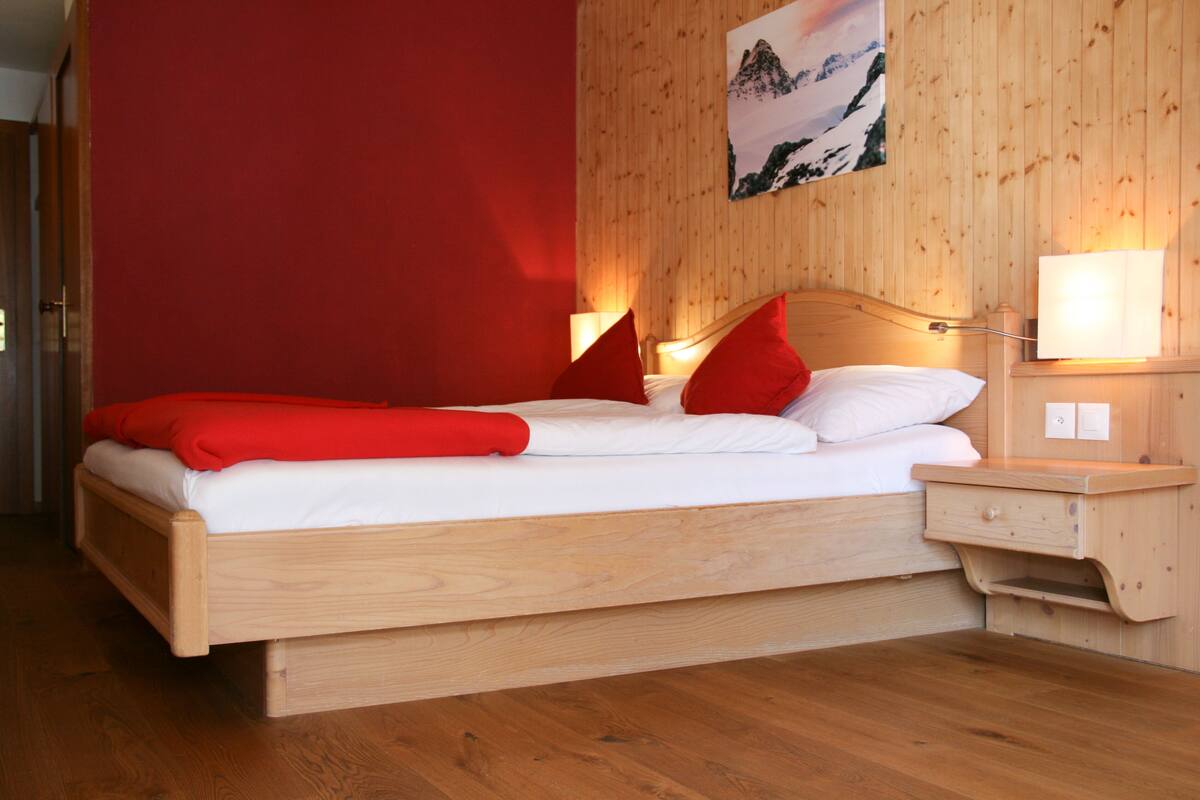 A wooden bed with red pillows and a white blanket, a nightstand with a lamp, and a wooden wall with a picture.